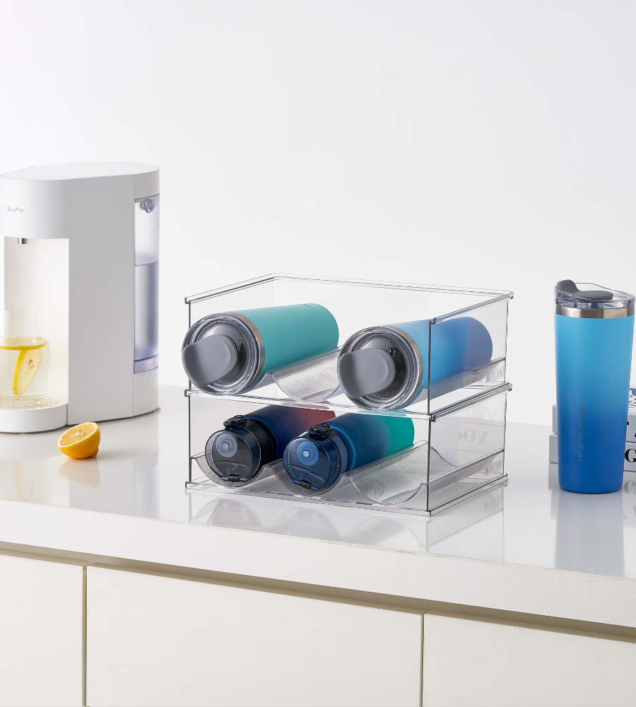 Der BNB x VCN Flaschenhalter KLAR - Set 2. Stk hält vier wiederverwendbare Flaschen auf einer weißen Küchentheke organisiert, während ein blauer Reisebecher, eine halb geschnittene Zitrone und ein weißer Getränkebereiter mit Zitronenwasser in der Nähe stehen.