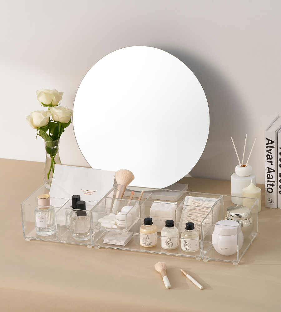 Ein Waschtisch mit rundem Spiegel, weißen Rosen in einer Vase, Hautpflege und Make-up im BNB x VCN SLIDE Schubladen Organizer KLAR - HOCH (verschiedene Größen), Pinsel, Wattepads, Diffusorstäbchen und Bücher auf der rechten Seite - alles auf einer beigen Fläche.