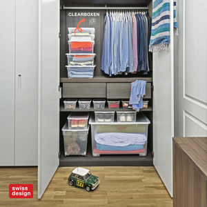 Ein organisierter Kleiderschrank mit der Rotho Aufbewahrungsbox CLEAR 46L (55x37,5x32cm), hängenden Hemden, gefalteter Kleidung und Schubladen. Auf dem Holzboden steht ein Spielzeugauto. Der Schrank ist mit einem CLEARBOXEN-Etikett und einem Schweizer Design-Logo in der Ecke versehen.