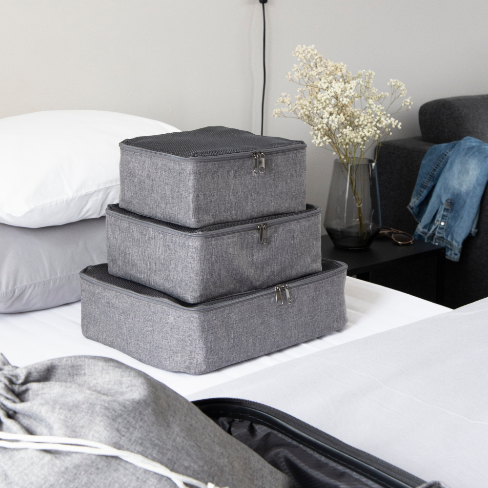 Drei Bigso TRAVEL Organizer S Würfel in Grau liegen gestapelt auf einem ordentlich gemachten Bett. Auf dem Nachttisch liegen Trockenblumen, Brillen, Jeans und Teile eines Kissens und Koffers - ideal für alle, die Koffer organisieren wollen.