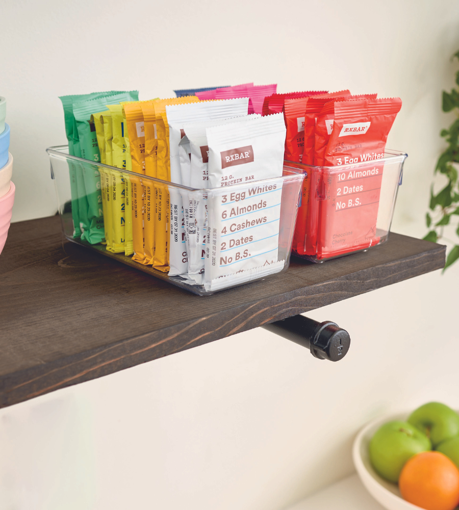 Un organiseur de tiroir iDesign LINUS HAUT contient des barres protéinées RXBAR colorées sur une étagère en bois foncé, avec un bol de fruits en dessous.
