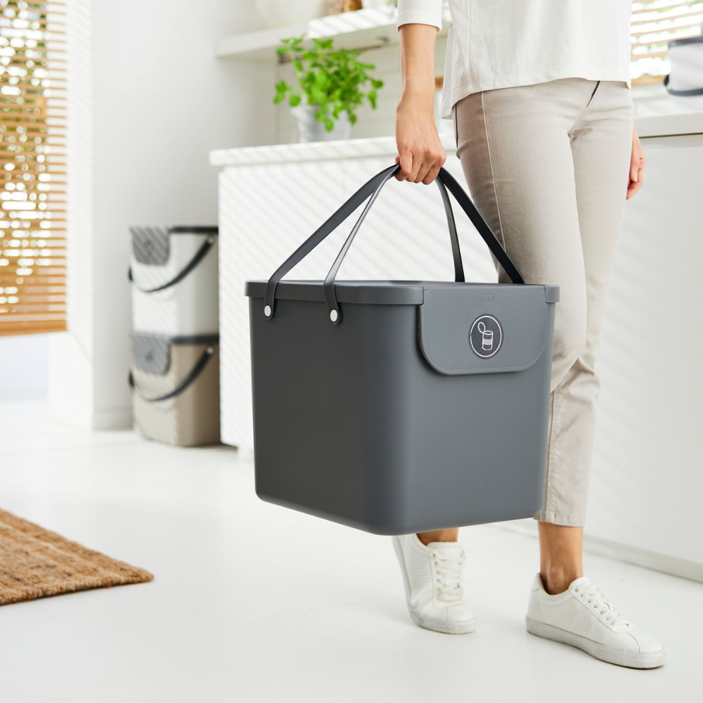 Eine Person in heller Kleidung und weißen Turnschuhen trägt ein großes Rotho ALBULA Recycling Müllsystem 40L - Anthrazit mit Griffen in einer hellen, modernen Küche. Im Hintergrund sind Pflanzen und Küchenutensilien zu sehen.