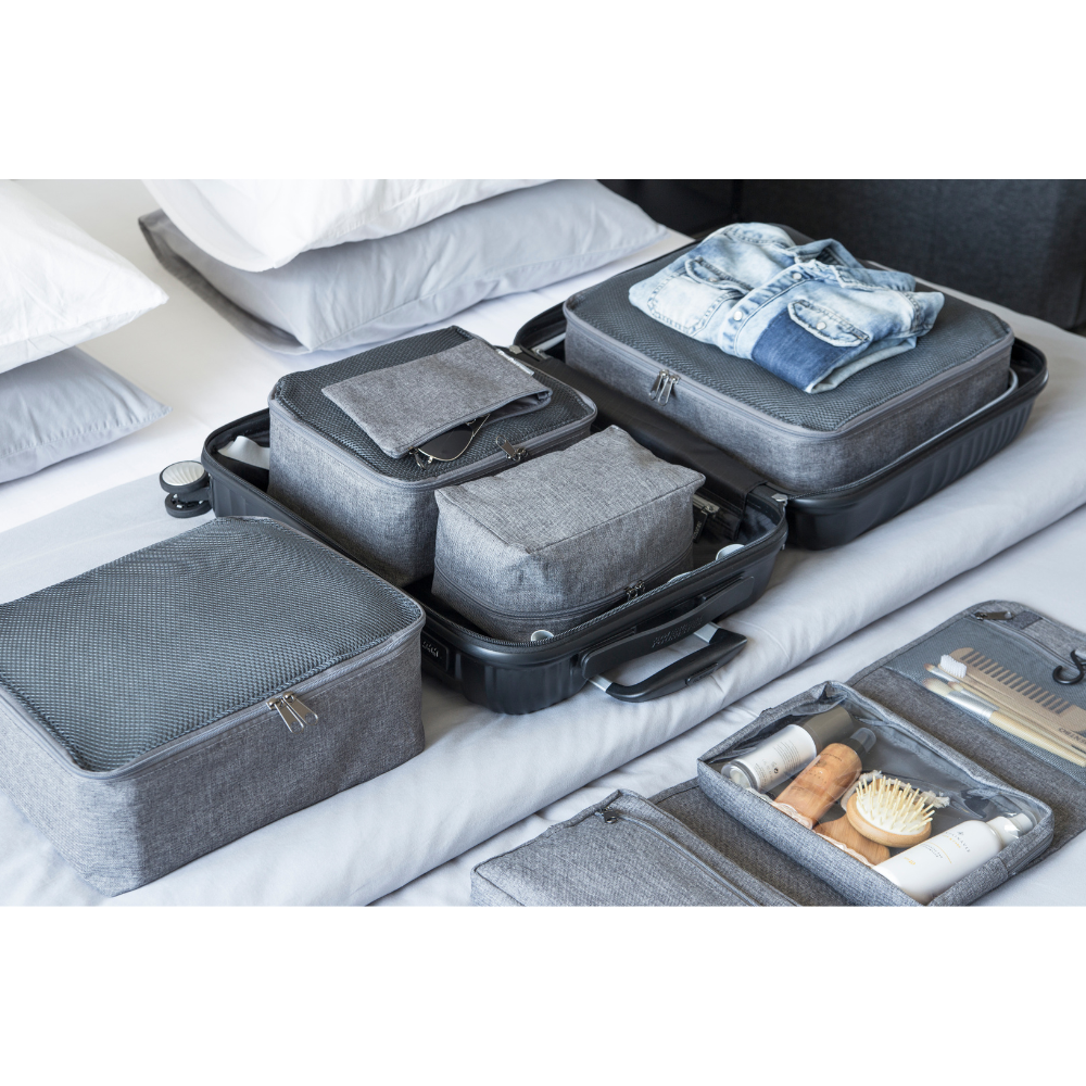 Ein ordentlich gepackter offener Koffer mit organisierten grauen Packwürfeln und einem Jeanshemd liegt auf einem Bett neben dem Bigso TRAVEL Organizer S in Grau, der Pflege- und Toilettenartikel aufnimmt.