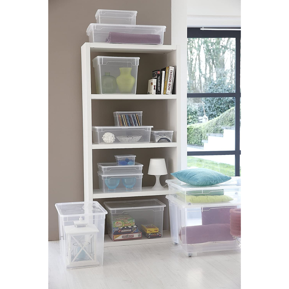 In einem weißen Regal stehen Bücher, Vasen, eine Lampe und eine Rotho Aufbewahrungsbox CLEAR 5L (33,5x19x11cm) mit verschiedenen Gegenständen. Weitere Rotho-Boxen - einige gefüllt mit Kissen oder Spielzeug - sind auf dem Boden neben einem großen Fenster gestapelt.