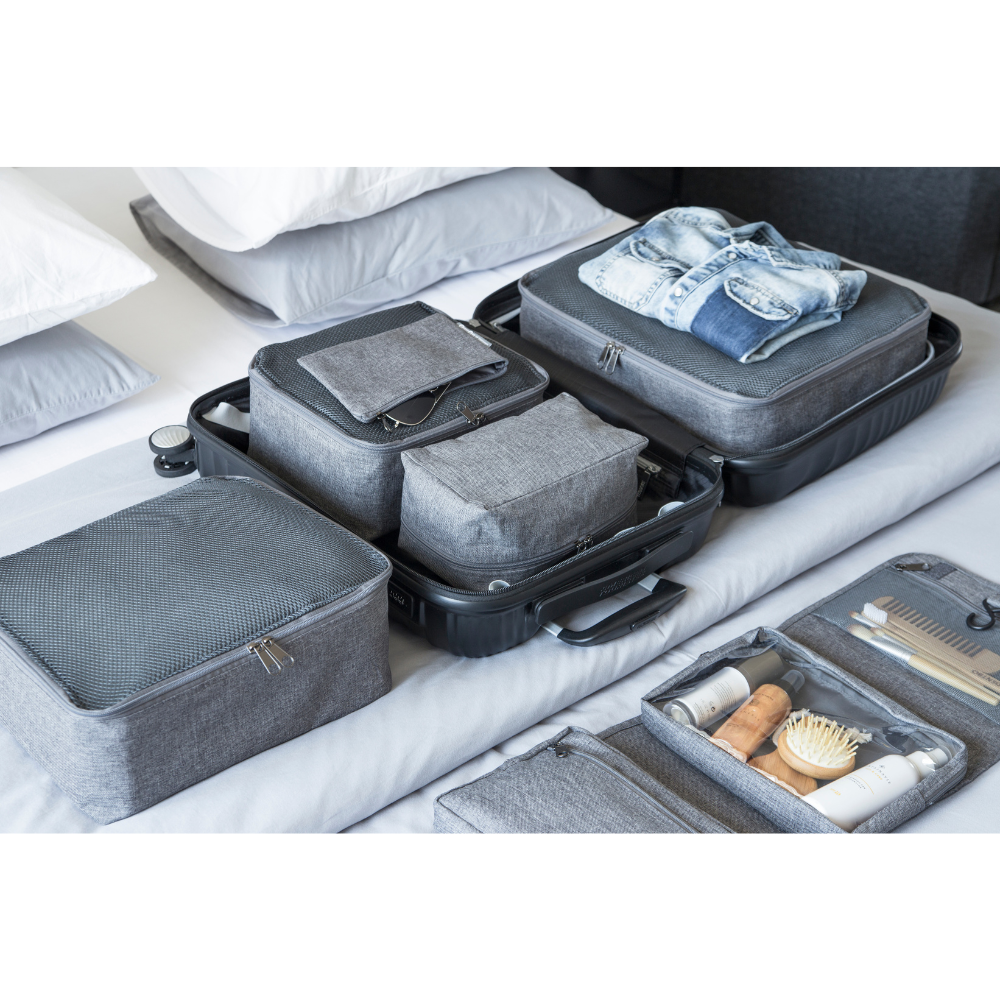 Ein ordentlich organisierter offener Koffer mit dem TRAVEL Organizer L - Grau von Bigso und Taschen sowie Kleidung ist umgeben von Toilettenartikeln in Reisegröße, einem Kamm und einer Bürste - alles auf einem Bett mit weißen Kissen im Hintergrund.