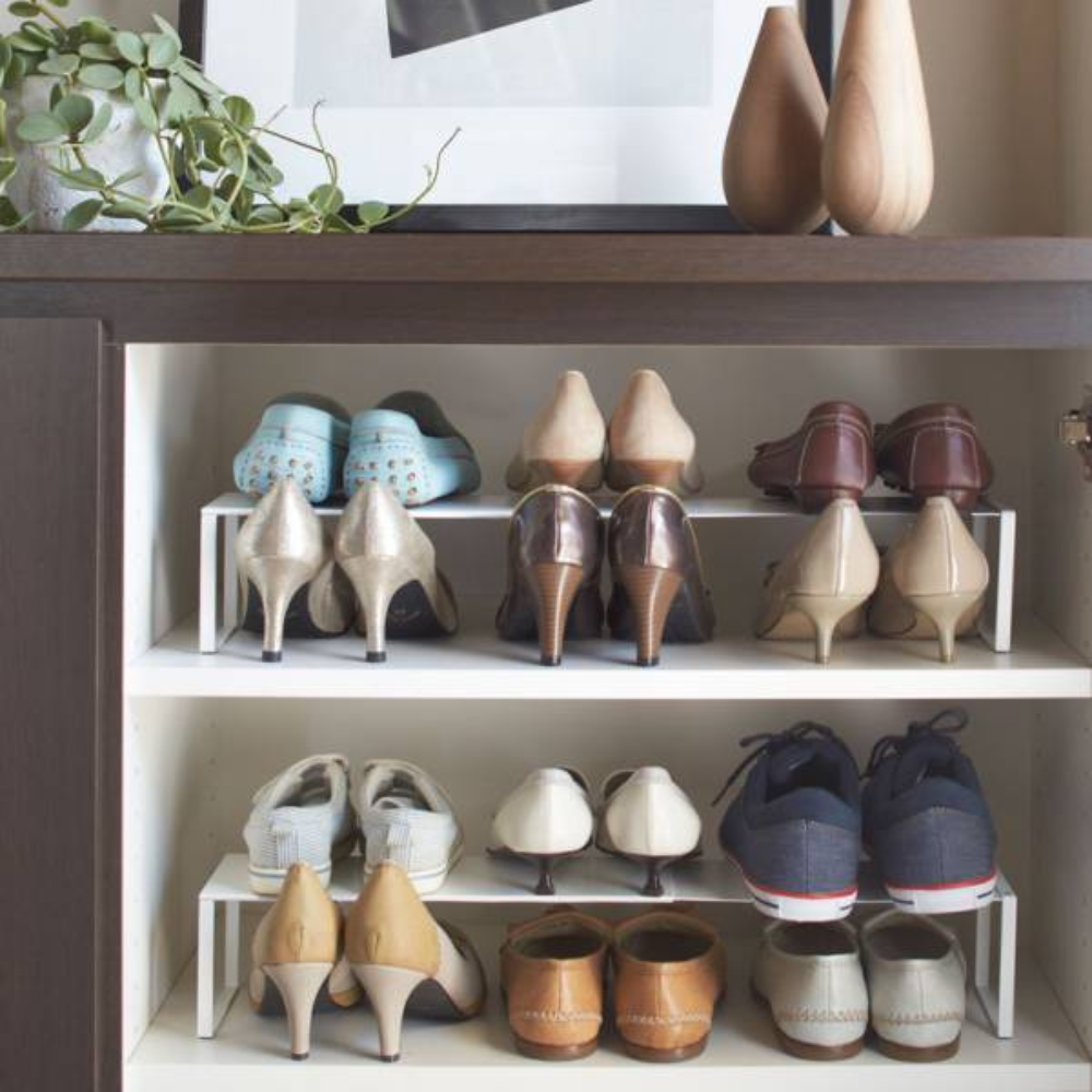 Der FRAME Schuhregal ausziehbar - Weiss von YAMAZAKI hält High Heels, Flats und Sneaker auf weißen Böden unter einem dunklen Holzschrank; oben setzen Vasen, Pflanzen und Kunst elegante Akzente für cleveren Stauraum.