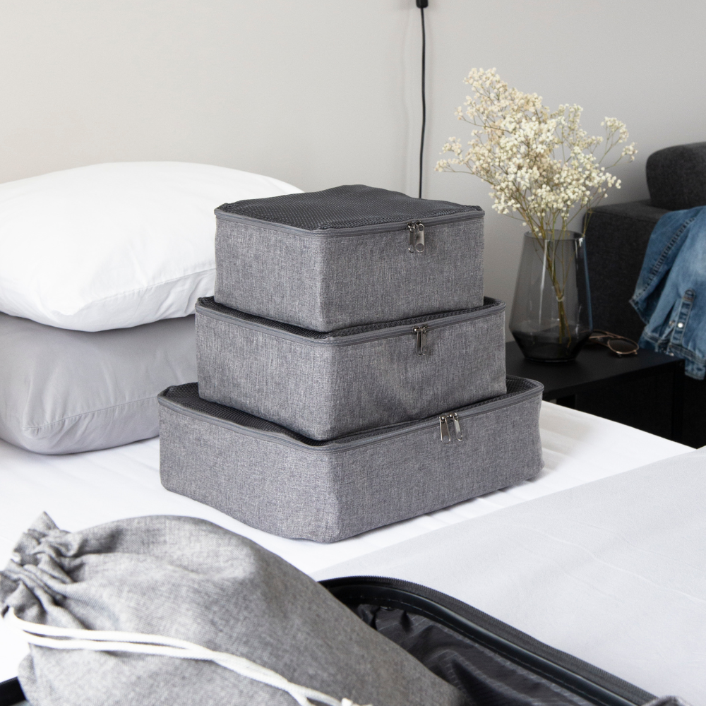 Drei Bigso TRAVEL Organizer L - Grau Packwürfel liegen gestapelt auf einem aufgeräumten Bett. Ein passender grauer Kordelzugbeutel und ein offener Koffer stehen in der Nähe, mit einem Nachttisch und einer Vase mit getrockneten Blumen im Hintergrund.