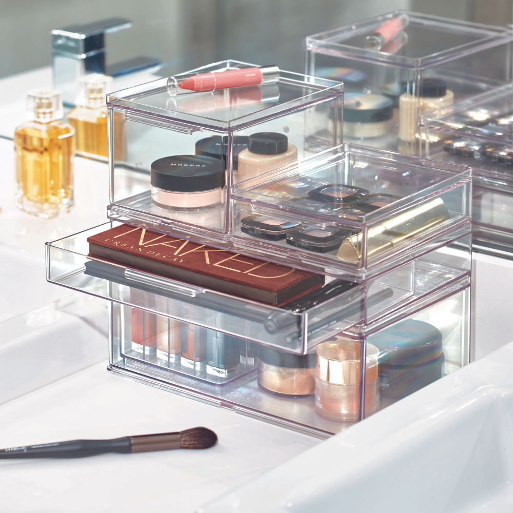 Modular HOME EDIT organizer with drawer by iDesign, in various sizes, filled with palettes, foundation, lipstick, and brushes, stands next to perfume bottles and a makeup brush on the white bathroom counter.