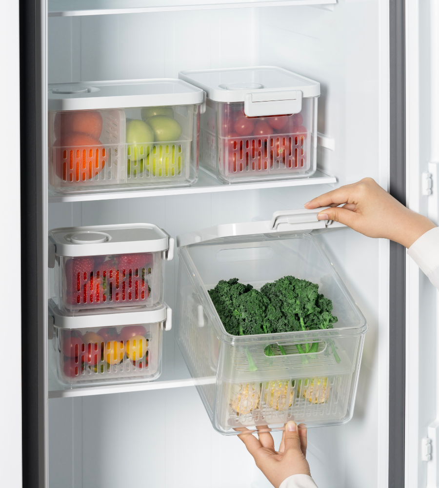 Eine Person bewahrt grünes Blattgemüse in einer durchsichtigen DATEMATE Frischaltebox KLAR Weiss mit Haltbarkeits-Timer von BNB x VCN im Kühlschrank auf. Dank ihres stapelbaren Designs ordnen ähnliche Boxen Obst und Gemüse übersichtlich im Regal.