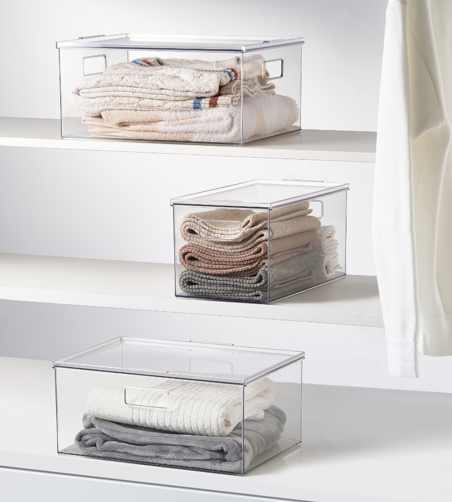 Trois boîtes de rangement empilables avec couvercle KLAR de BNB x VCN, extra longues et de différentes tailles, sont posées sur des étagères blanches, remplies de vêtements ; à droite, un vêtement blanc est suspendu au bord.