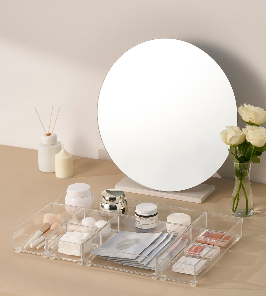 Ein runder Spiegel steht auf einer beigen Fläche hinter einem SLIDE Schubladen Organizer KLAR von BNB x VCN, der mit Hautpflegetiegeln, Schminkpinseln, Gesichtsmasken und Rouge gefüllt ist. Weiße Rosen, Kerzen und ein Schilfrohrdiffusor sind in der Nähe angeordnet.