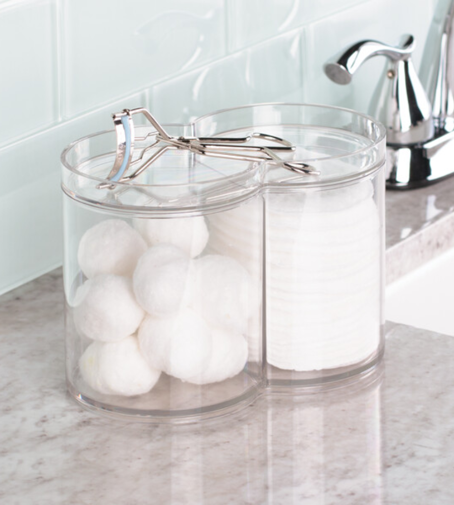 Der CLARITY Kosmetik Organizer KLAR von iDesign steht befüllt mit Wattebällchen und Pads auf dem Bad-Tresen. Eine Wimpernzange liegt auf dem Deckel, Wasserhahn und geflieste Rückwand erscheinen im Hintergrund.