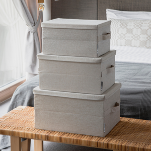 Drei beige SOFT Aufbewahrungsboxen von Bigso (35x26x19cm) sind auf einer geflochtenen Bank in einem Schlafzimmer gestapelt. Diese stilvollen, zusammenklappbaren Stoffaufbewahrungsboxen mit Deckel bieten praktische und elegante Aufbewahrungslösungen.