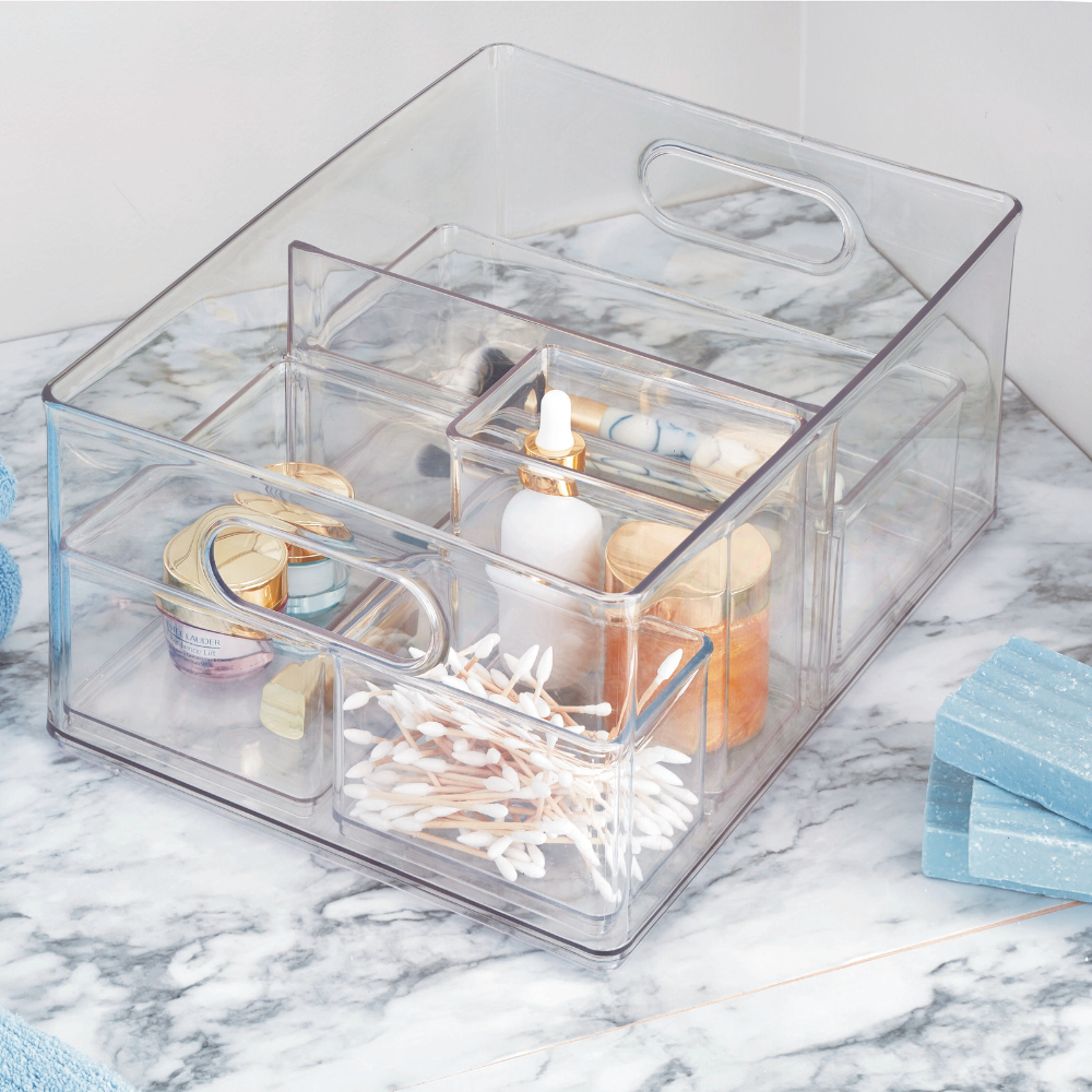 Der iDesign HOME EDIT Aufbewahrungsbehälter KLAR mit 2 Fächern (25.4x34.3x15.2cm) organisiert Wattestäbchen, Tropfflaschen und Kosmetik auf der Marmorplatte neben blauen Handtüchern und Seifen - stapelbar, herausnehmbare Trennwand.