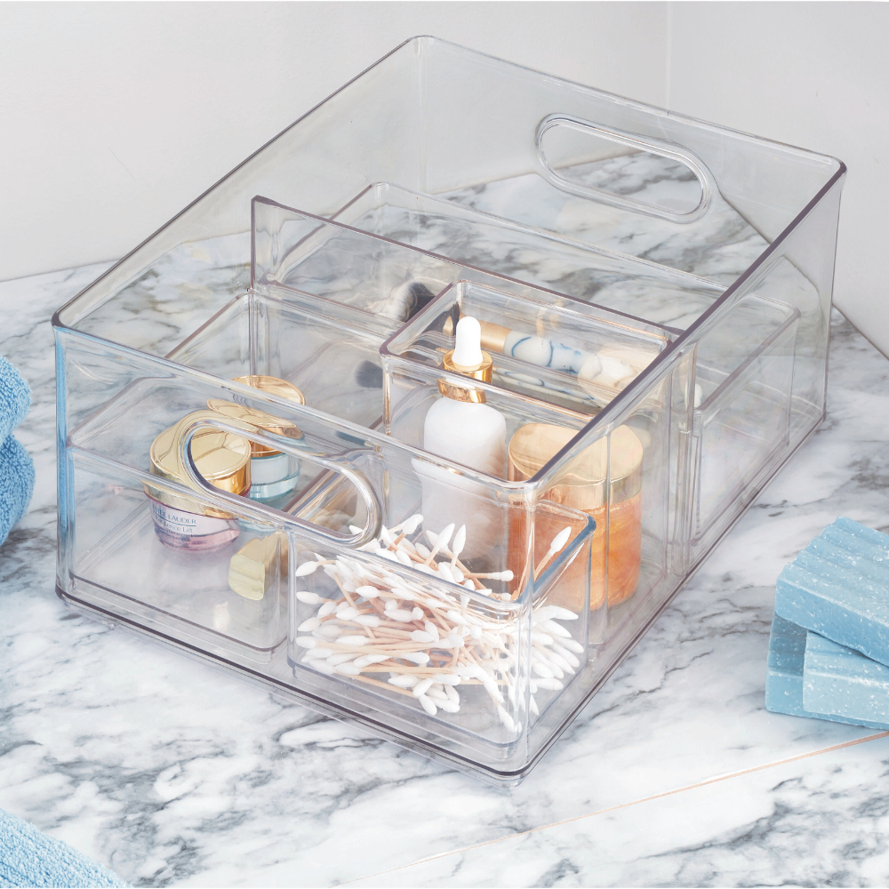 Transparent organizer with cosmetic products and cotton swabs on a marble table, next to towels and soap.