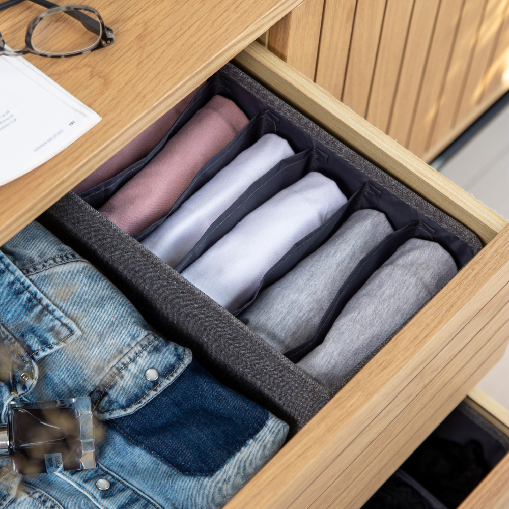 Eine offene Schublade mit dem Bigso SOFT Organizer Grau - 6 Fächer schmal präsentiert farblich sortierte Hemden, eine Jeansjacke und Parfüm. Gläser und Papier liegen oben auf der Schublade und veranschaulichen clevere Aufbewahrungsideen.