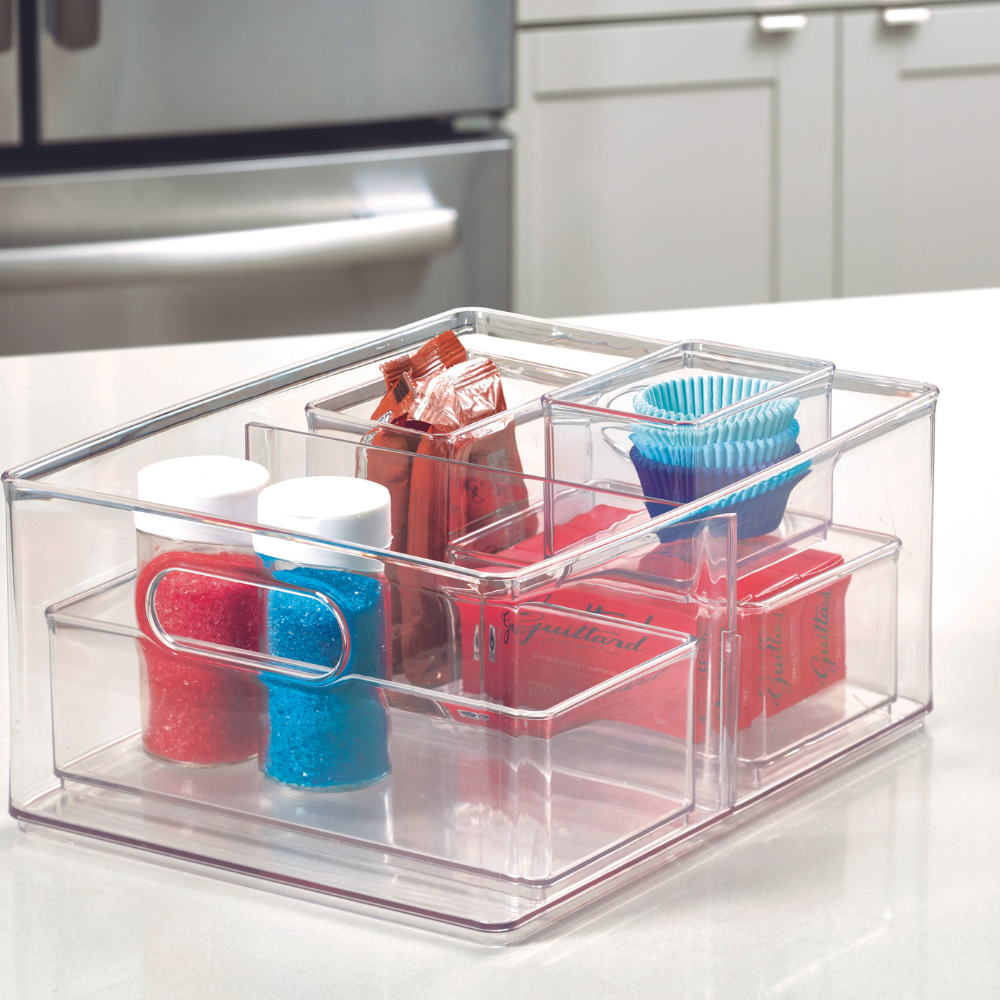 Ein HOME EDIT - Aufbewahrungsbehälter KLAR mit 2 Fächern von iDesign (25.4x34.3x15.2cm) organisiert Kuchenmischungen, Cupcake-Förmchen, Toppings und Streusel ordentlich auf der Küchenplatte; im Hintergrund sind Kühlschrank und weiße Schränke zu sehen.