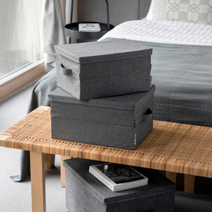 Zwei SOFT Aufbewahrungsboxen Grau L 45x34x25cm von Bigso sind auf einer geflochtenen Bank am Fußende eines Bettes gestapelt. Eine kleinere Box und ein Buch liegen daneben, alles sanft beleuchtet vom natürlichen Licht des Fensters.