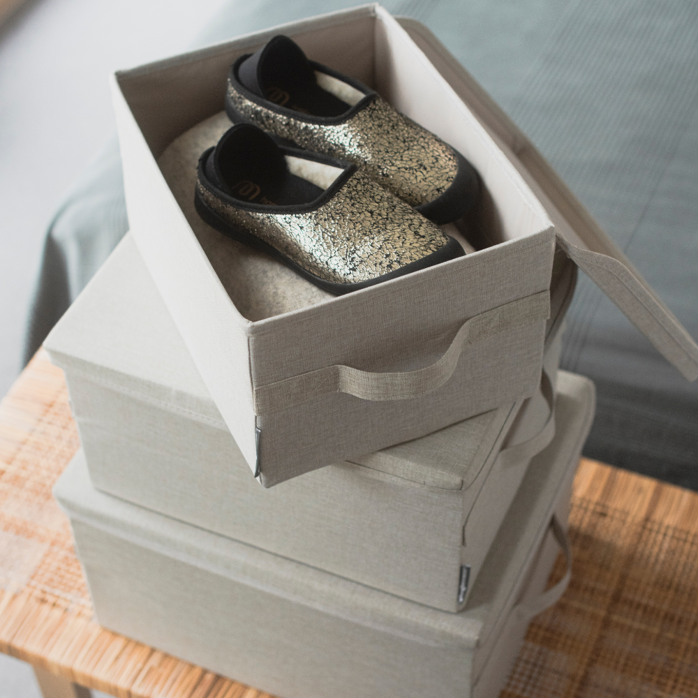 Ein Paar schwarz-goldene Krabbelschuhe sitzt in einer offenen, zusammenklappbaren Box auf zwei geschlossenen, stapelbaren SOFT Aufbewahrungsbox Beige L - 45x34x25cm von Bigso auf einer Holzfläche neben einem Bett.