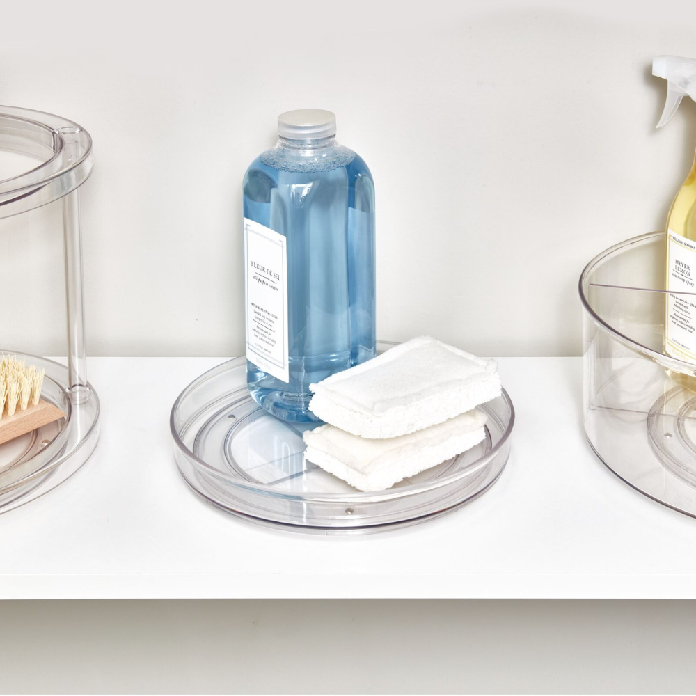 Ein durchsichtiger iDesign HOME EDIT - Drehteller Lazy Susan (23x3,5cm) hält eine blaue Flasche und zwei weiße Schwämme auf einem weißen Regal, in dessen Nähe weitere Behälter und Reinigungsmittel sichtbar sind.