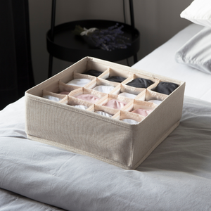Der SOFT Organizer Beige - 16 Fächer von Bigso aus Stoff bietet perfekte Aufbewahrung für Kleidung in verschiedenen Farben. Er steht auf einem Bett mit hellgrauer Bettwäsche, daneben ein schwarzer Beistelltisch und Lavendel-Deko.