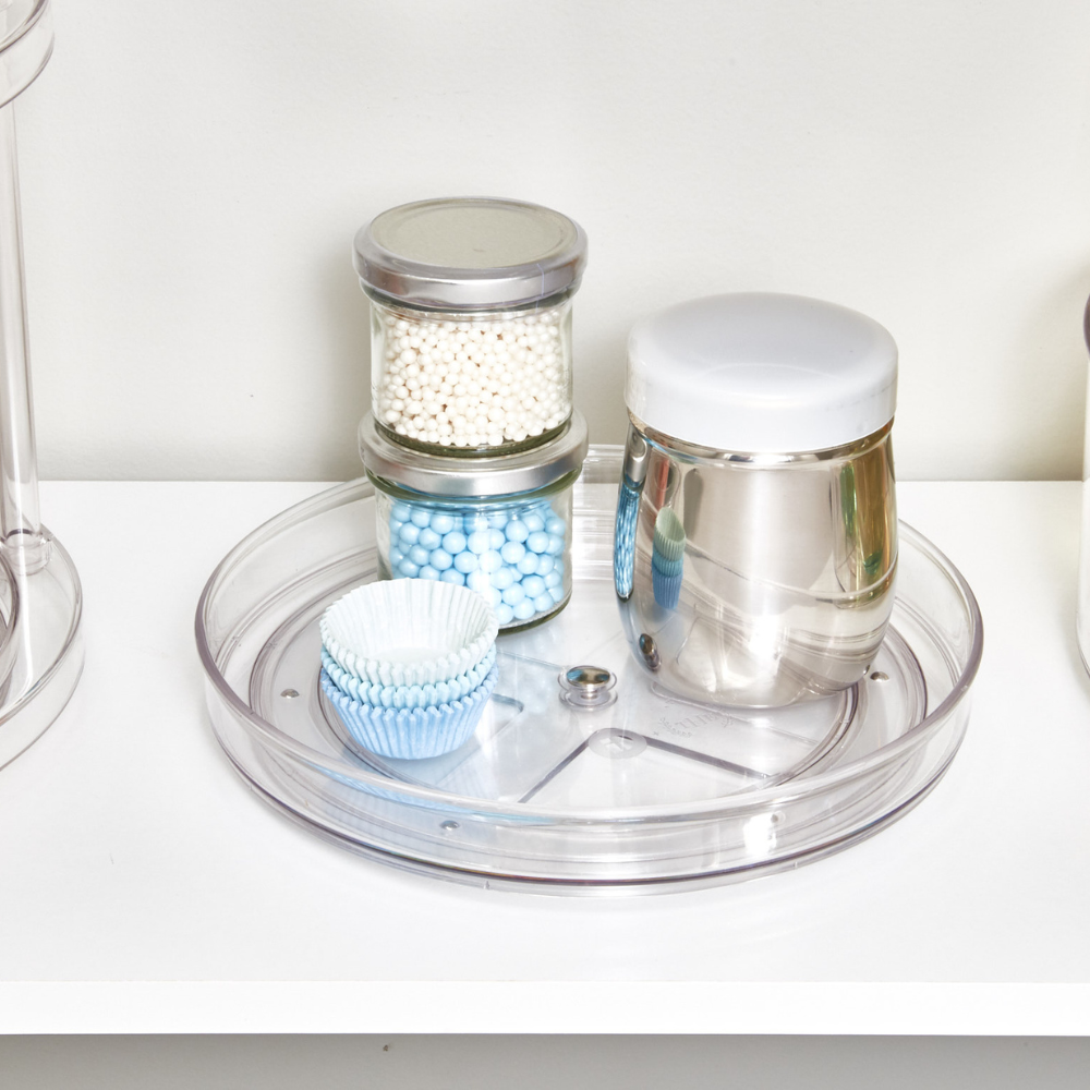 Der iDesign HOME EDIT - Drehteller Lazy Susan KLAR (23x3,5cm) ordnet Gläser mit weißen und blauen Wattestäbchen, einen Edelstahlbehälter mit weißem Deckel und hellblaue Papierförmchen für Muffins auf einer weißen Ablage.