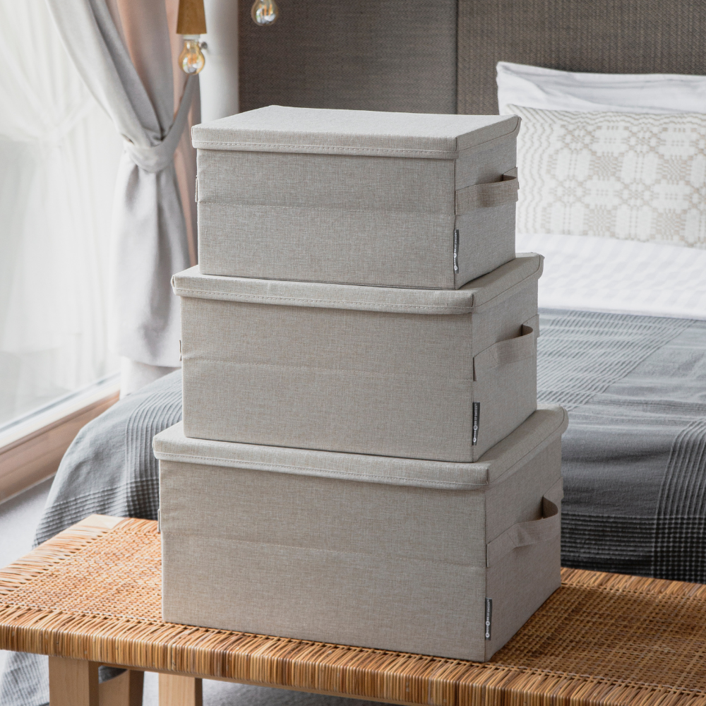 Drei Bigso SOFT Aufbewahrungsboxen in Beige (Größe M, 40x30x22 cm) sind im Schlafzimmer auf einer Bank gestapelt; im Hintergrund sieht man ein ordentlich gemachtes Bett und ein Fenster mit transparenten Vorhängen.