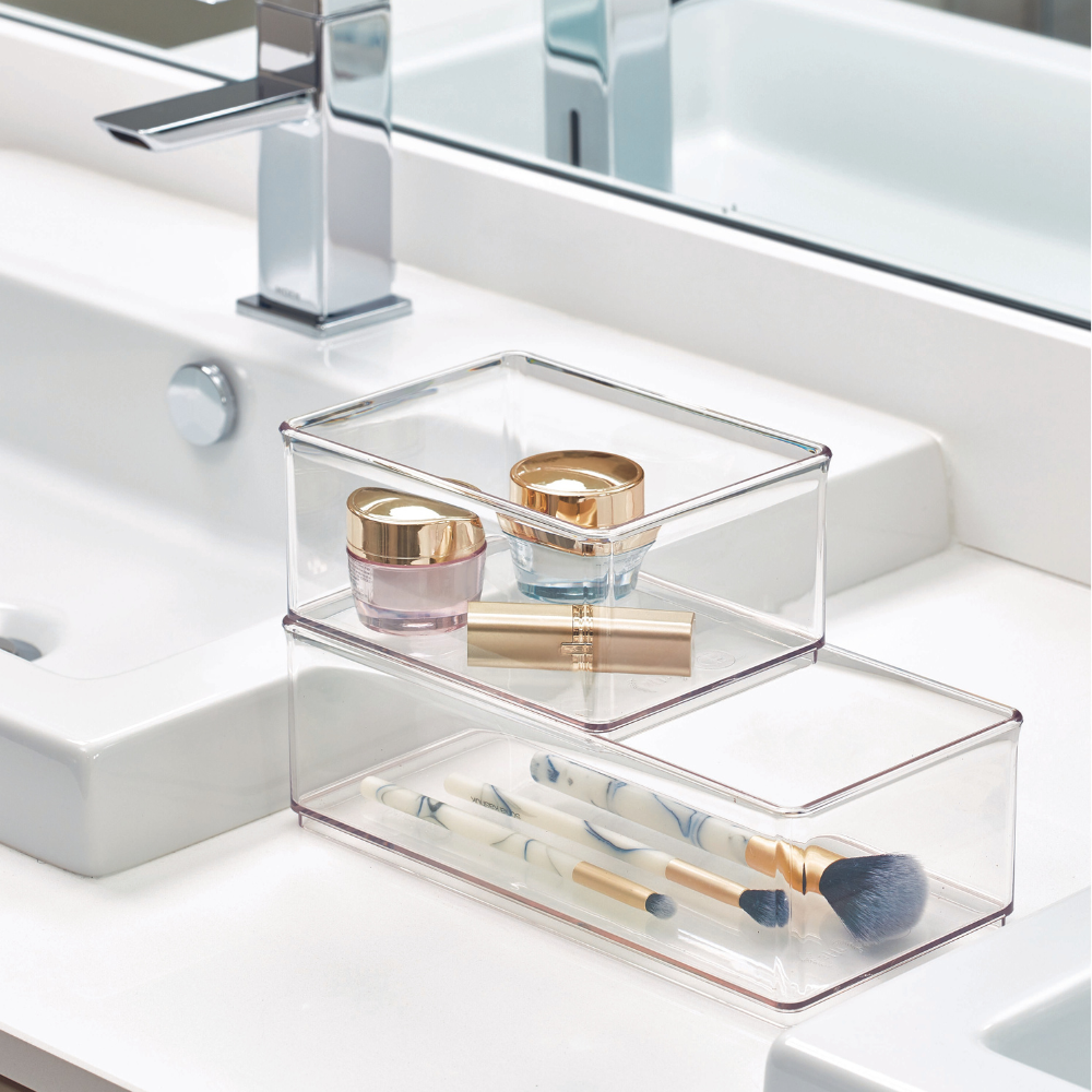 Transparent storage boxes with cosmetic products and brushes on a white sink next to the faucet and mirror.
