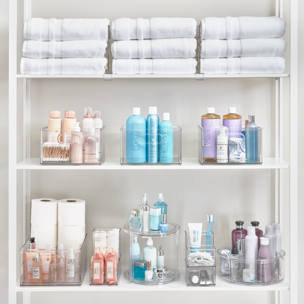 Ein weißes Regal zeigt gefaltete Handtücher und Toilettenartikel im iDesign HOME EDIT - Aufbewahrungsbehälter KLAR mit 2 Fächern (25,4x34,3x15,2cm) und bietet stilvolle, stapelbare Aufbewahrung mit herausnehmbarem Trennwand für eine ordentliche Organisation.