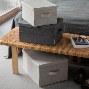 Drei Bigso SOFT Aufbewahrungsboxen Beige L (45x34x25 cm): Zwei sind auf eine geflochtene Holzbank gestapelt, eine steht darunter. Daneben liegen Magazin, Brille und Fernbedienung. Im Hintergrund befindet sich ein Bett mit karierter Decke.