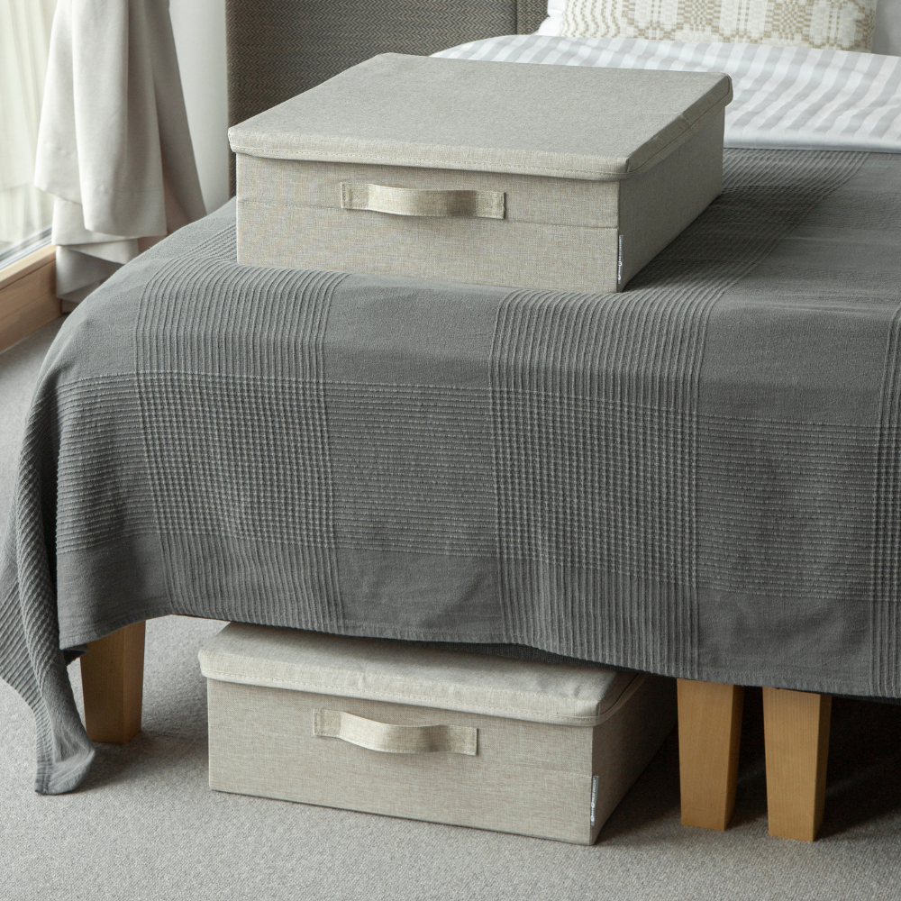 Zwei beige SOFT Aufbewahrungsboxen von Bigso, eine auf einer grau karierten Bettdecke und eine zur Unterbett-Lagerung teils unter dem Bett mit Holzfüßen in einem gemütlichen, mit Teppich ausgestatteten Raum.