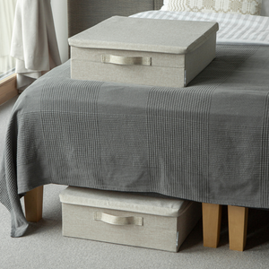 Zwei beige SOFT Aufbewahrungsboxen von Bigso, eine auf einer grau karierten Bettdecke und eine zur Unterbett-Lagerung teils unter dem Bett mit Holzfüßen in einem gemütlichen, mit Teppich ausgestatteten Raum.